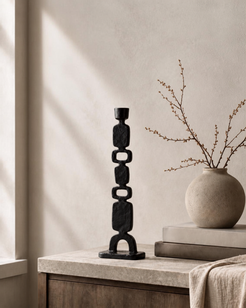 Handcrafted black decorative candlestick holder with stacked geometric rings, displayed on a stone-topped side table next to a beige ceramic vase with dried branches.
