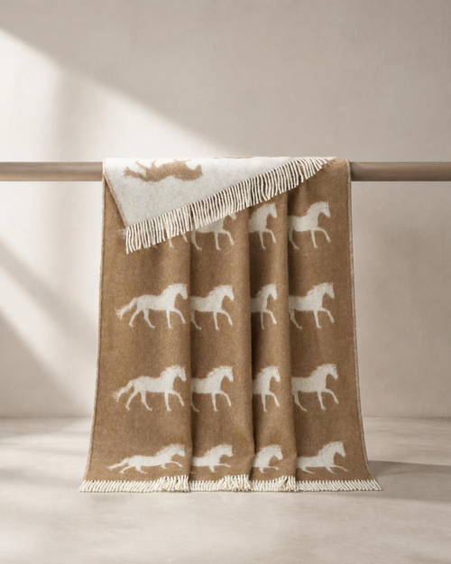 A beige and white woven throw blanket with a repeating horse pattern and fringed edges, draped over a horizontal wooden rod in a softly lit neutral interior.